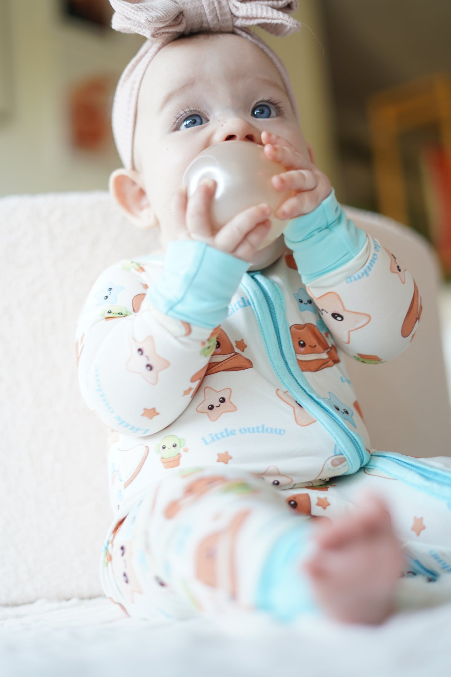 Close up of baby wearing our blue Cowboy Outlaw bamboo zippy romper with exclusive western print and pink bow headband sitting on a soft cream chair