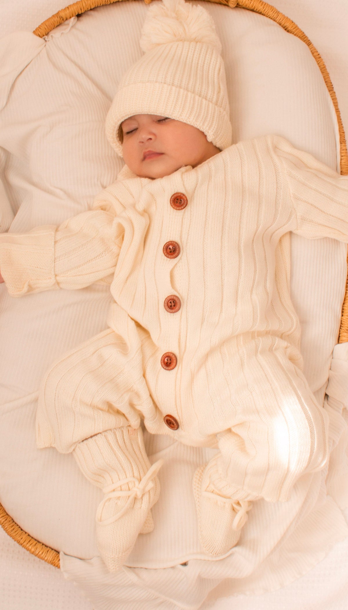 Newborn baby wearing the Vanilla Marshmallow knit bundle in a wicker bassinet with matching beanie and booties