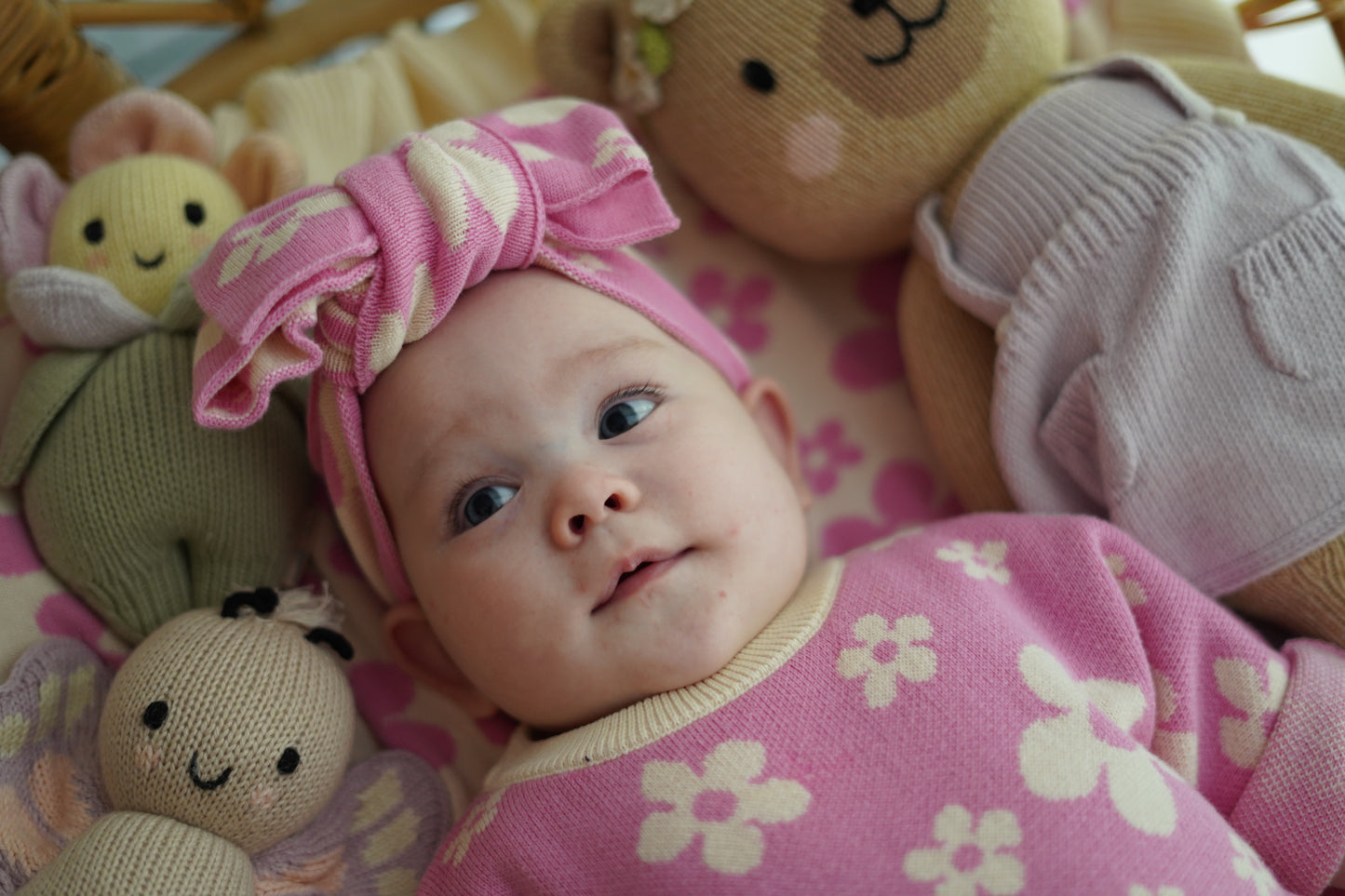 Baby wearing our Violet Knit Headband and matching pink floral knit outfit lying with soft plush toys in a cosy styled setting
