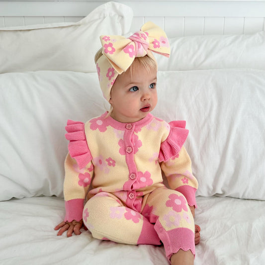 Baby girl wearing the Buttercup Princess Romper, a soft 100% cotton knit romper featuring a pink floral design, frill shoulders and matching bow headband.