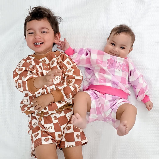 Smiling siblings wearing matching Little Violet Threads lounge sets, featuring the Atelier brown check set and pink boutique lounge set on a white background