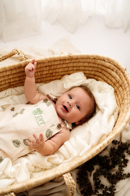 Baby lying in woven bassinet wearing Monstera Baby Overalls cotton leaf infant outfit