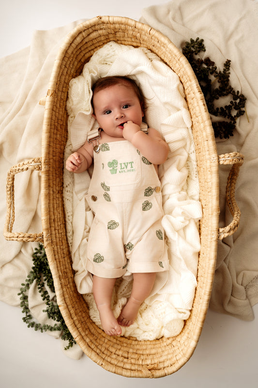 Baby lying in woven bassinet wearing Monstera Baby Overalls cotton leaf infant outfit