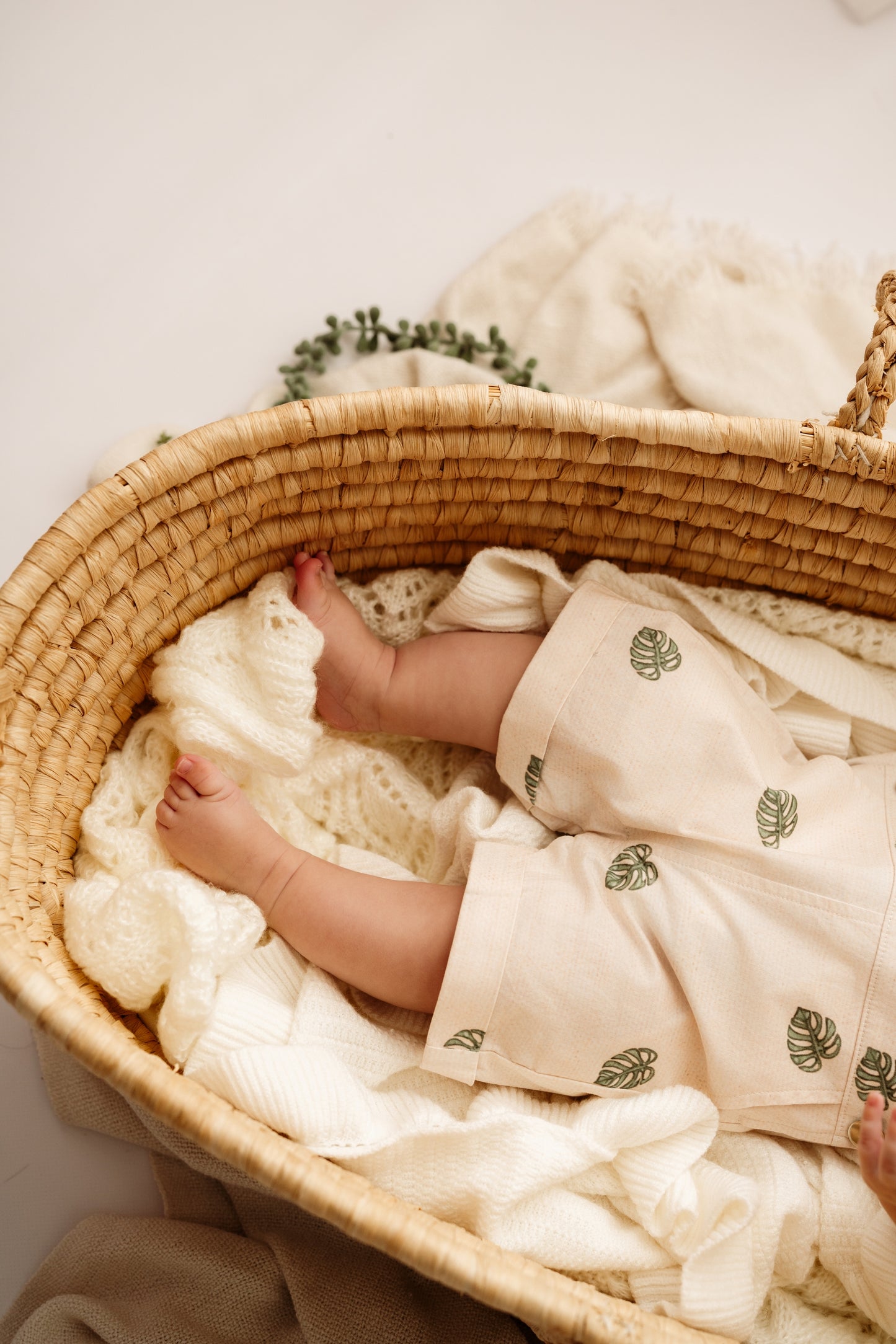 Baby legs in woven bassinet wearing Monstera Baby Overalls cotton leaf infant outfit