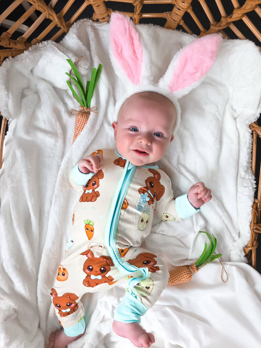 Baby wearing our Blue Bunny Bamboo Zippy Romper with bunny ears, lying on a soft white blanket with Easter carrot props