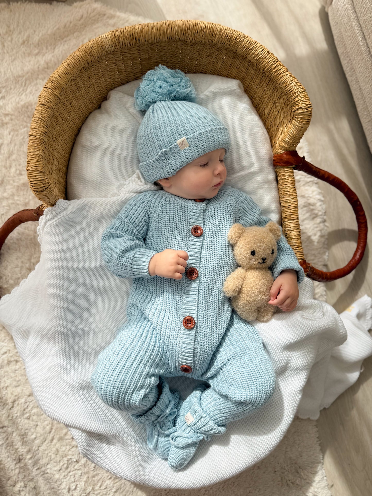 Baby wearing the Blue Haven Bundle soft blue cotton knit set with matching beanie and booties, lying in a wicker bassinet with a teddy bear