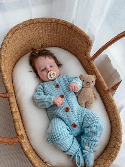 Baby wearing the Blue Haven Bundle soft blue cotton knit set with matching booties, lying in a wicker bassinet with a teddy bear
