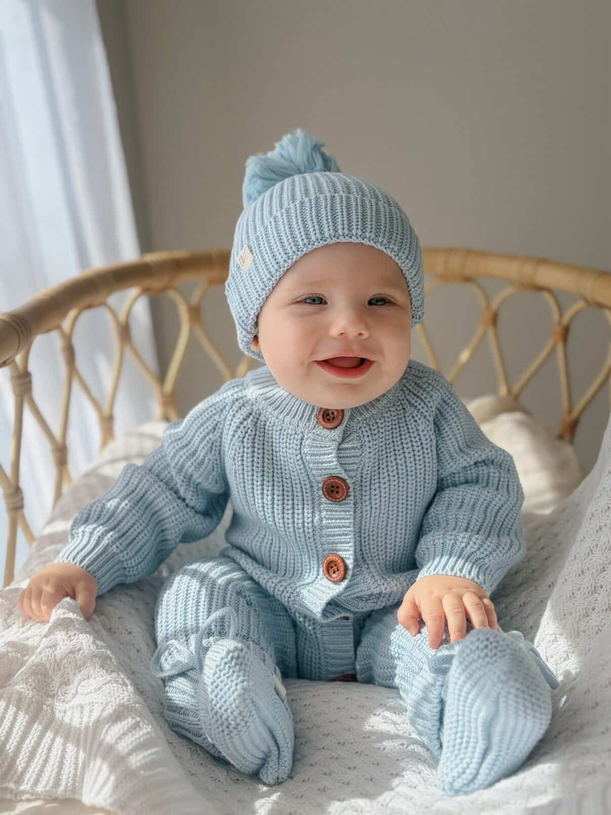 Baby wearing the Blue Haven Bundle soft blue cotton knit set with matching beanie and booties, sitting in a wicker chair