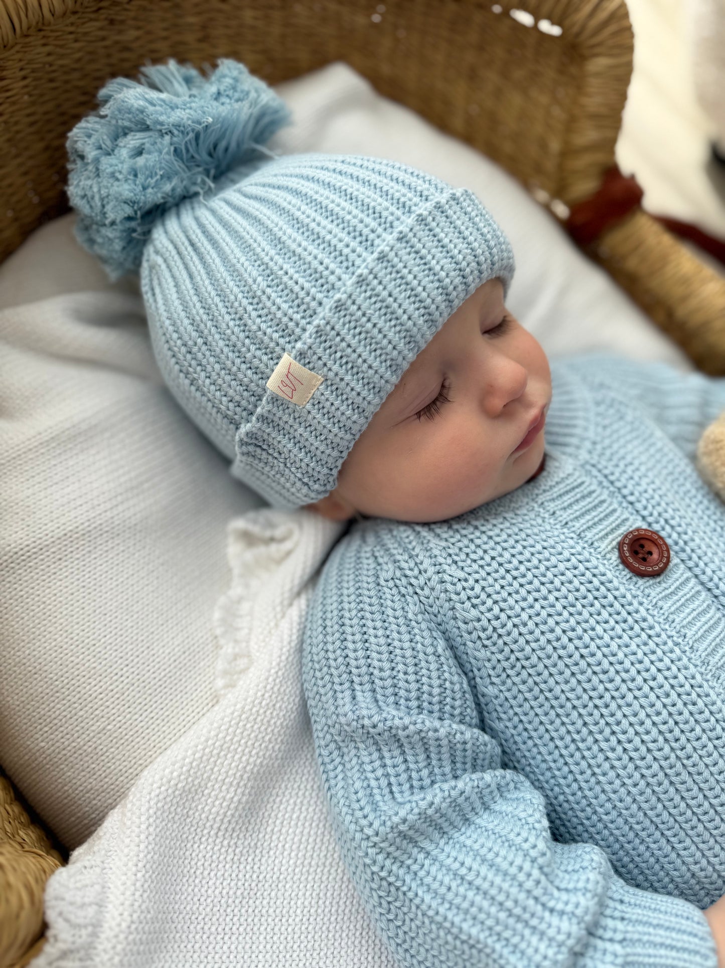 Blue Haven knit baby beanie with fluffy pom pom styled on sleeping baby in matching blue cotton knit outfit