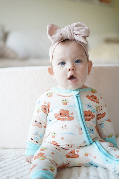 Baby wearing our blue Cowboy Outlaw bamboo zippy romper with exclusive western print and pink bow headband sitting on a soft cream blanke