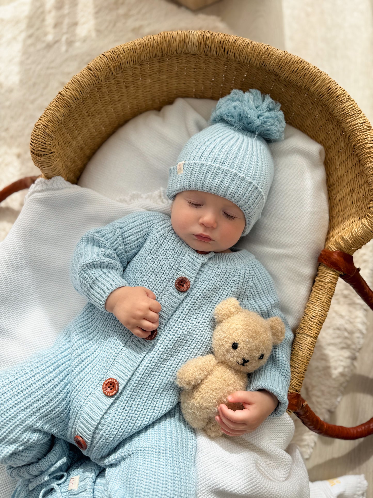 Baby wearing Blue Haven chunky knit romper set with matching blue beanie and booties, lying in wicker basket holding teddy bear
