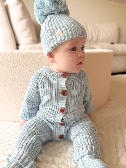 Baby wearing Blue Haven chunky knit romper with matching blue beanie sitting on a soft neutral lounge chair