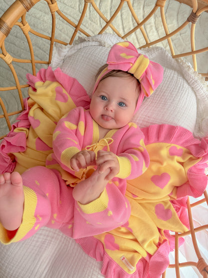 Baby girl wearing the pink and yellow Buttercup Princess cotton knit set with matching bow headband, lying on a heart knit blanket.