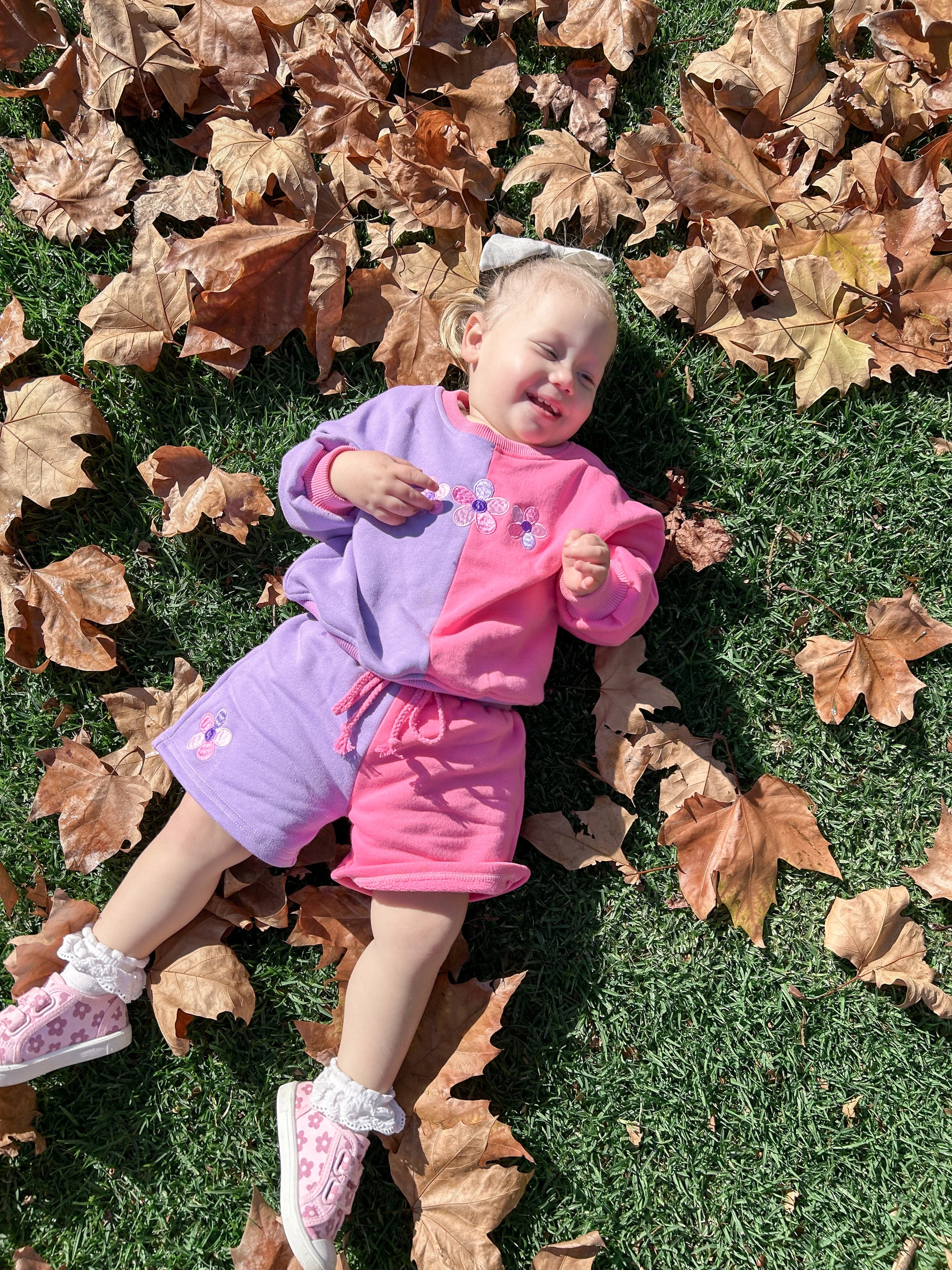 Toddler girl lying on autumn leaves wearing the Little Violet Threads La Dolce Lollie colourblock sweat set in pink and lilac