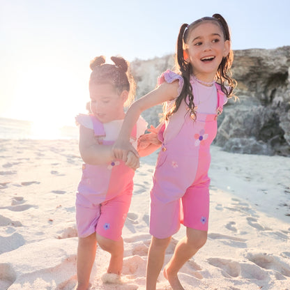 Girls wearing La Dolce Lollie pink floral overalls by Little Violet Threads on a sandy beach.