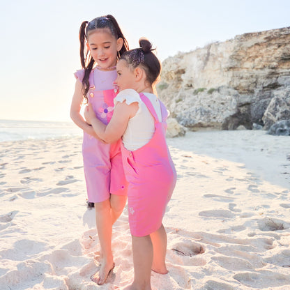 Two girls wearing La Dolce Lollie pink overalls by Little Violet Threads on the beach.