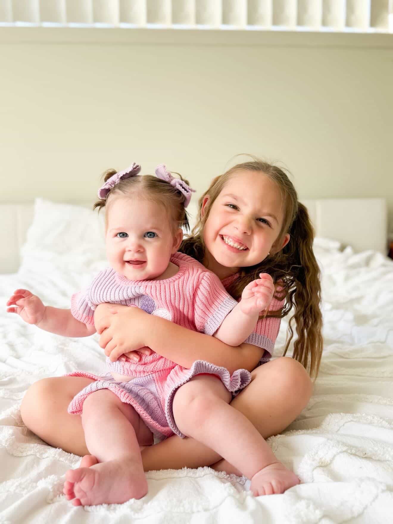 Two young sisters sitting on a bed wearing matching pink and lilac boutique knit outfits from Little Violet Threads La Dolce Lollie collection