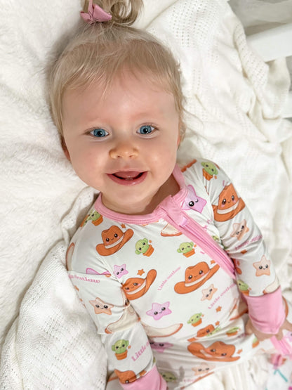 Baby girl wearing Little Outlaw bamboo zippy romper with pink western print lying on a soft white blanket