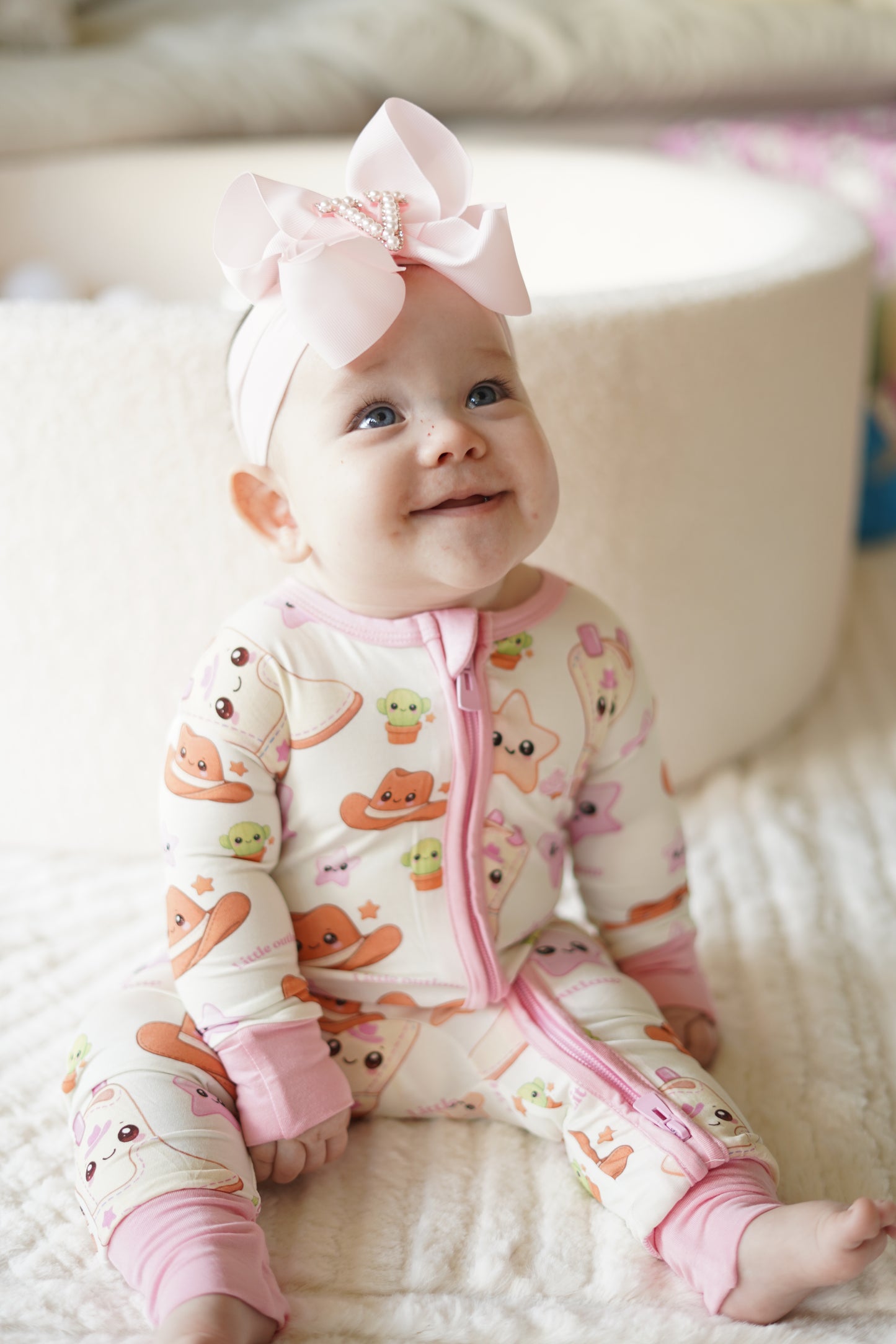 Baby girl wearing Little Outlaw pink bamboo zippy romper sitting on a soft neutral blanket with oversized pink bow headband