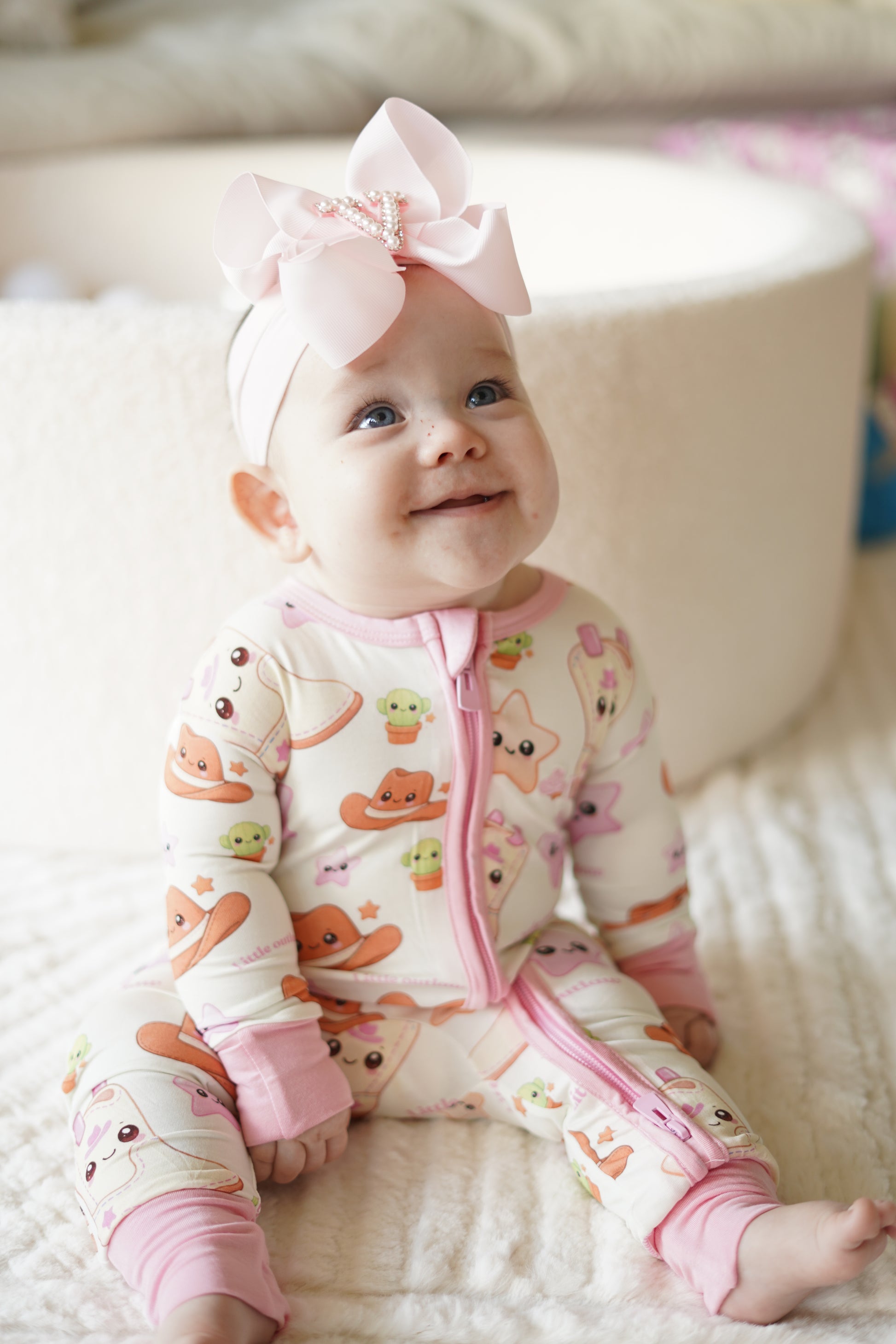 Baby girl wearing Little Outlaw pink bamboo zippy romper sitting on a soft neutral blanket with oversized pink bow headband
