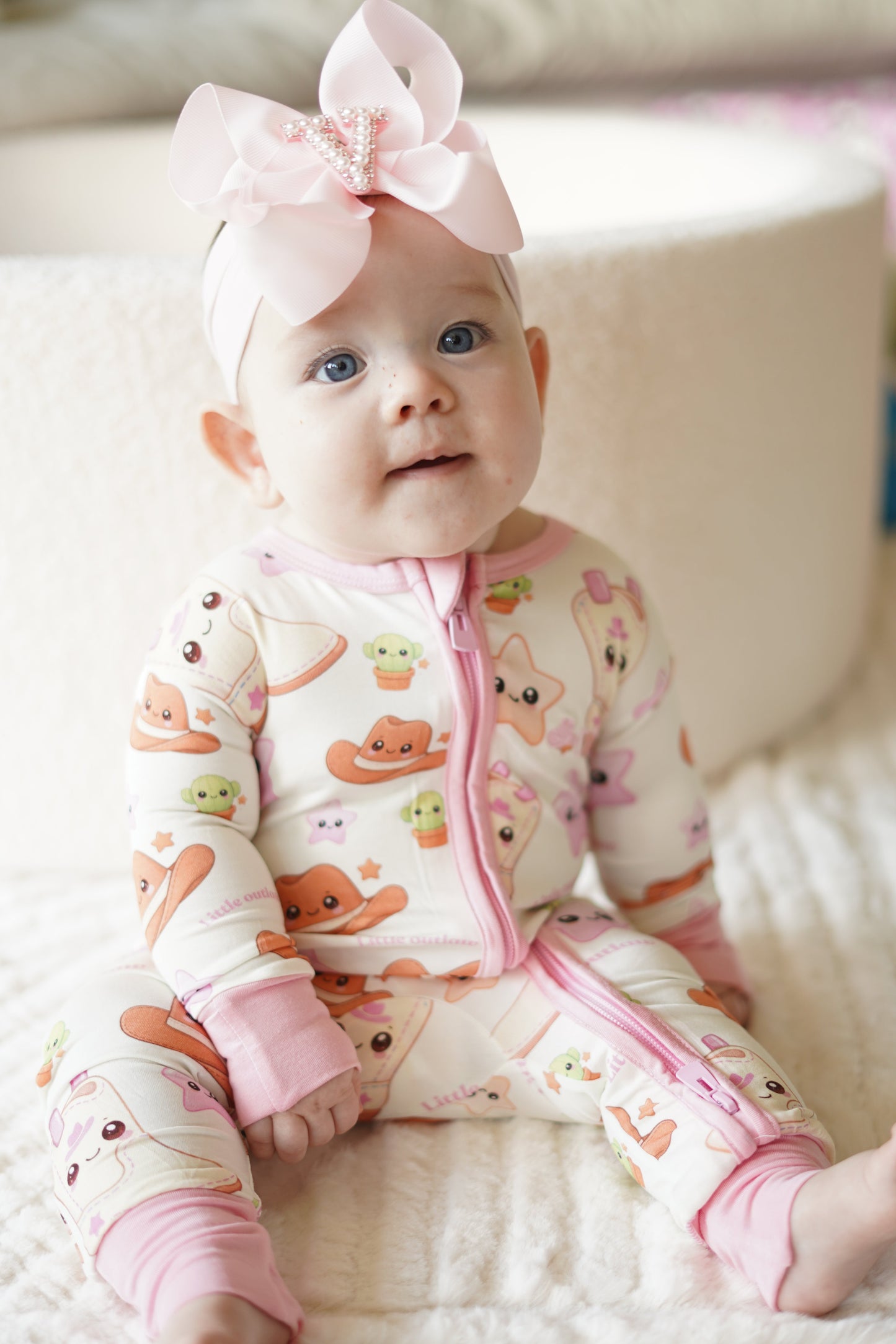 Baby girl wearing Little Outlaw pink bamboo zippy romper with oversized pink bow headband sitting on a soft neutral blanket