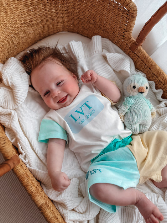 Baby wearing Little Violet Threads Pastel Maison lounge set with oversized tee and blue yellow colour block shorts in wicker bassinet