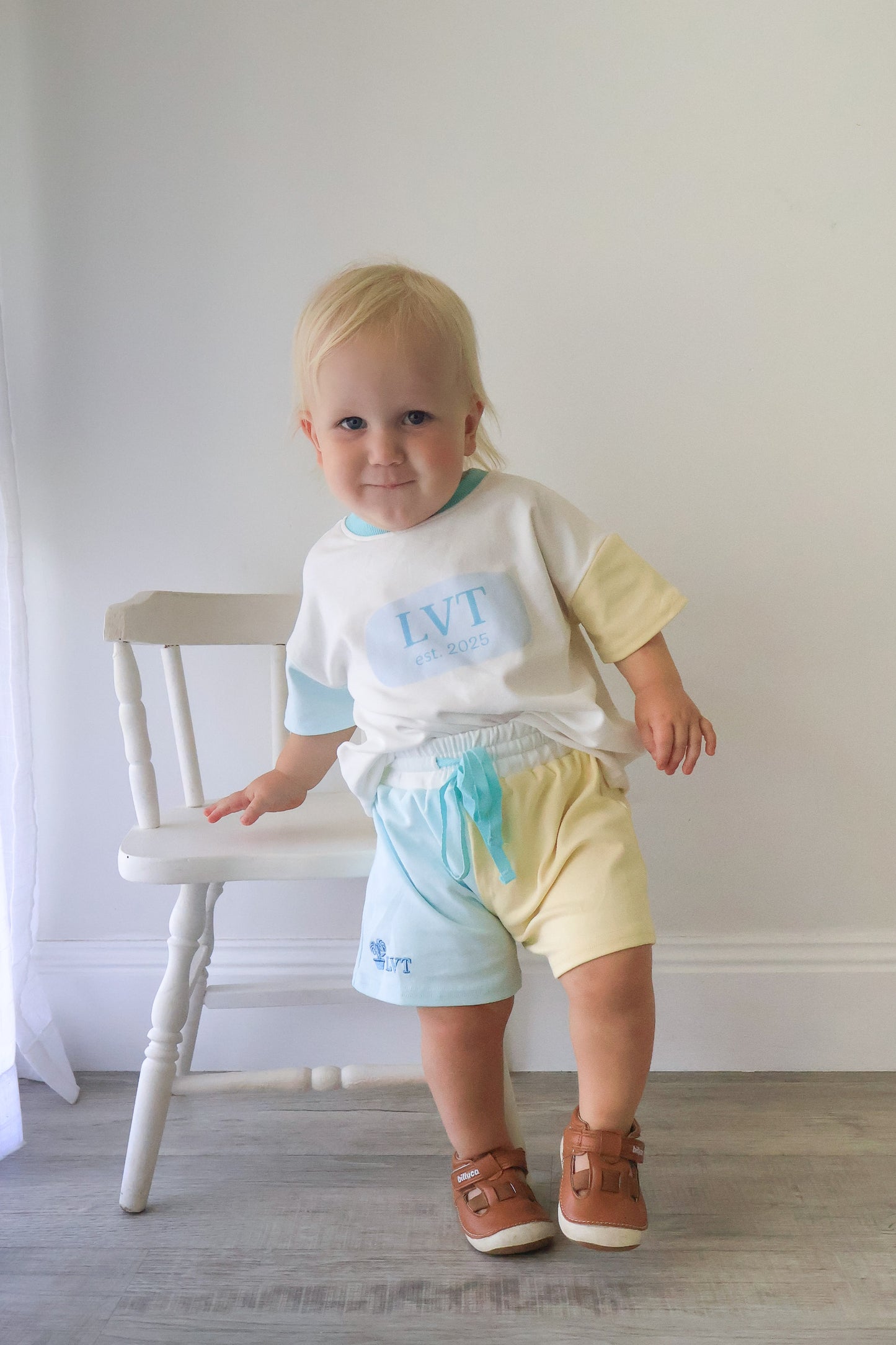 Toddler wearing Little Violet Threads Pastel Maison blue and yellow lounge set with oversized tee and colour block shorts standing beside white chair 