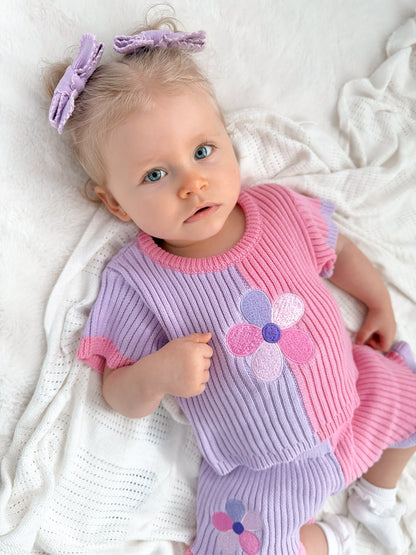Baby girl wearing a pink and lavender ribbed knit set with flower details and matching lavender bow hair clips.