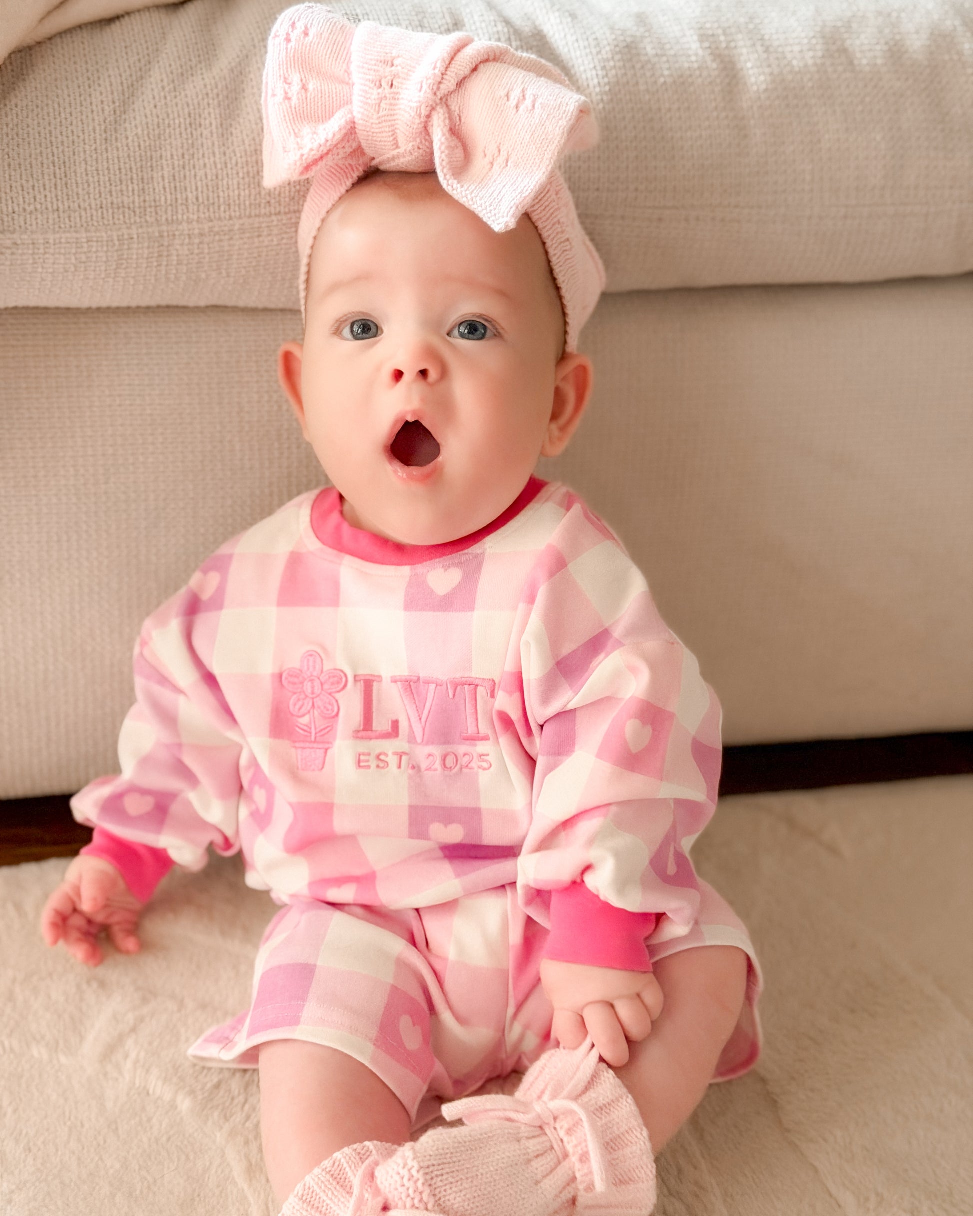 Baby girl wearing Little Violet Threads Pink Sugar Lounge Set in pink gingham sitting on lounge with matching oversized bow