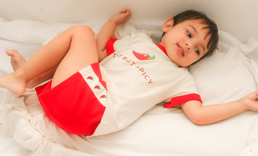 Child wearing our Tiny But Spicy matching kids set featuring oversized chilli graphic tee and red relaxed shorts lounging on soft cream bedding