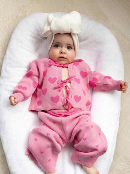 Baby wearing the Little Violet Threads Valentina Knit Set in pink heart print with matching oversized bow headband, lying on white cushion