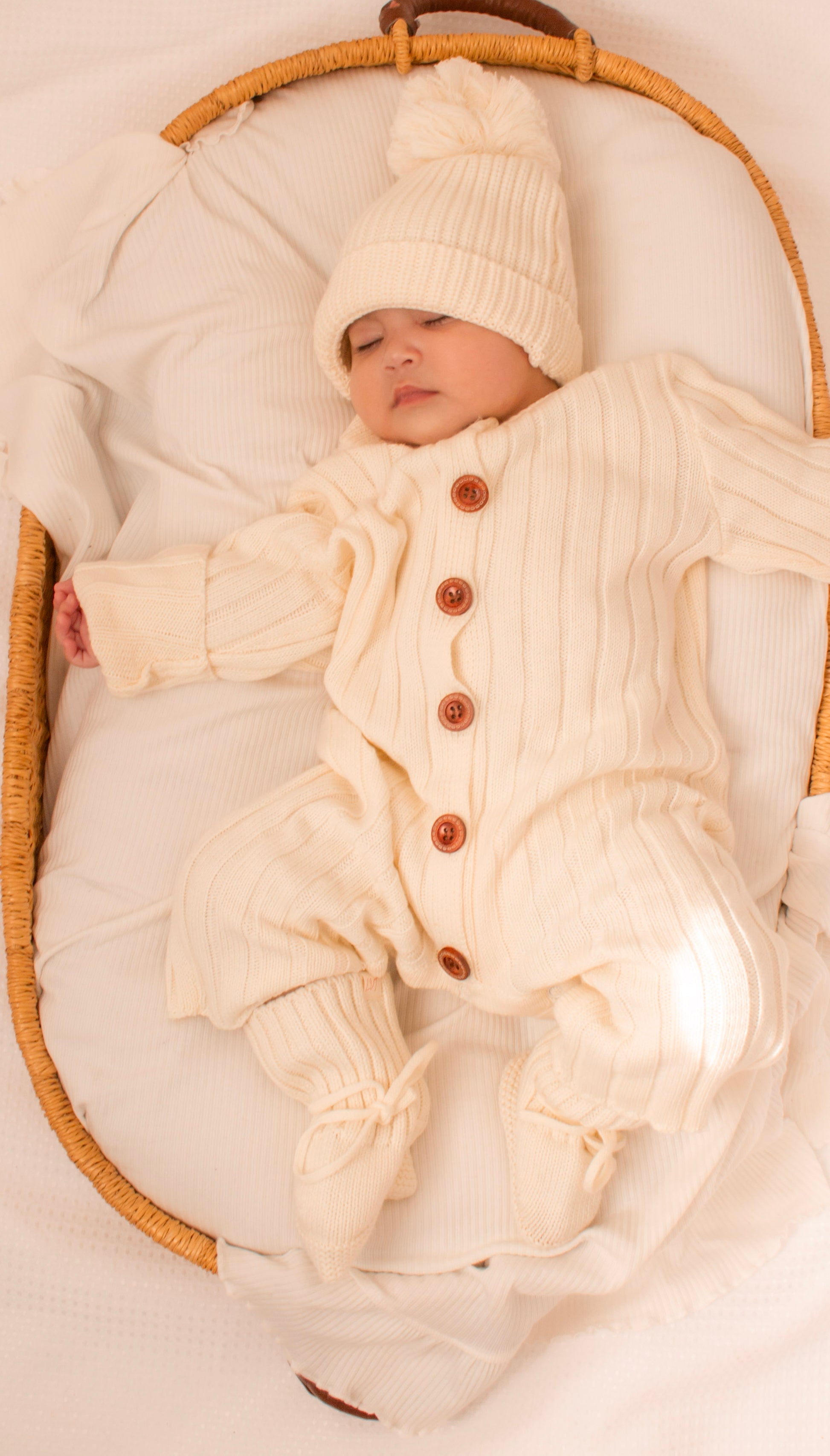 Newborn baby wearing the Vanilla Marshmallow knit bundle in a wicker bassinet with matching beanie and booties