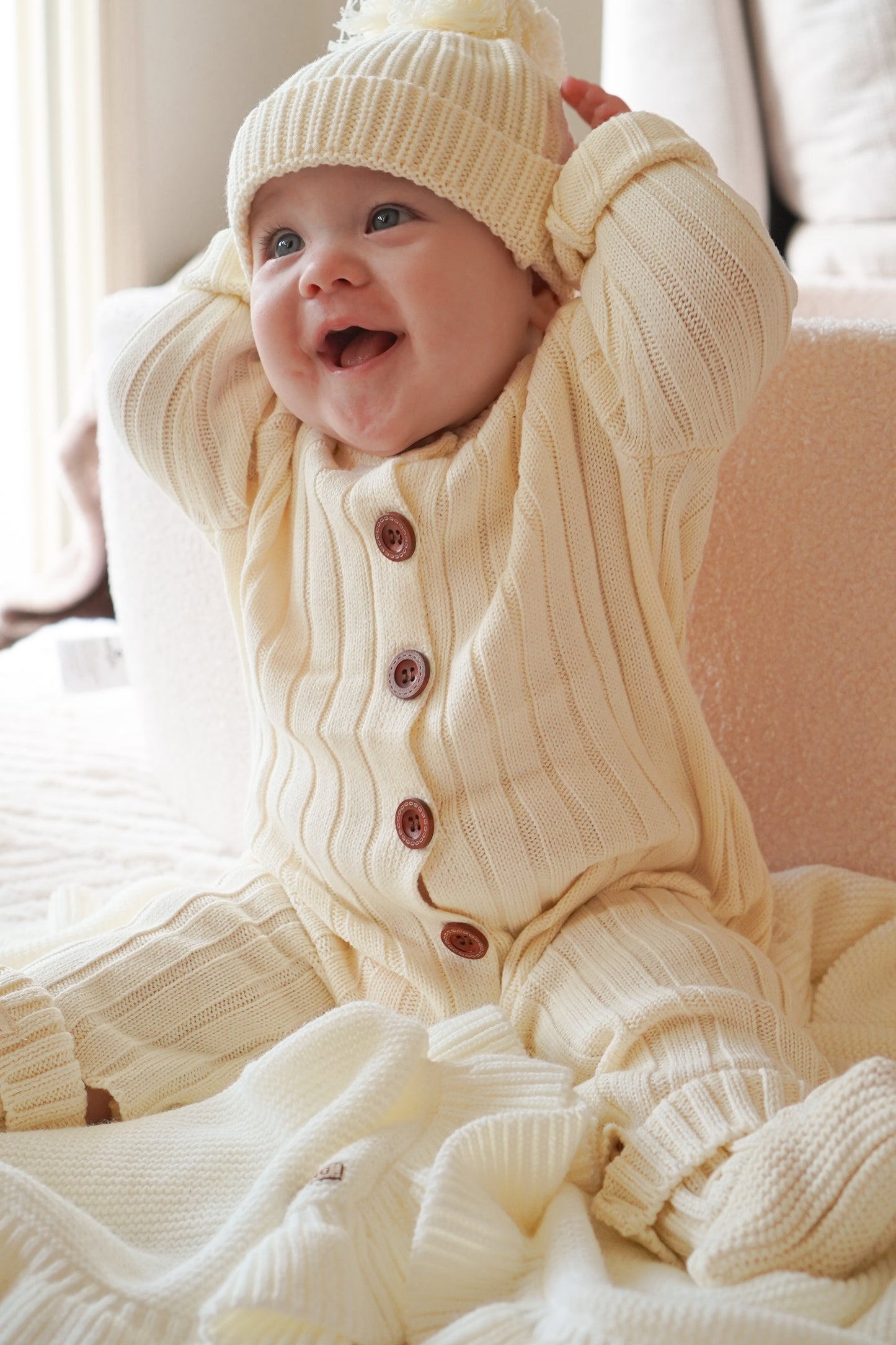 Smiling baby wearing the Vanilla Marshmallow knit bundle with matching beanie and booties sitting on a soft blanket