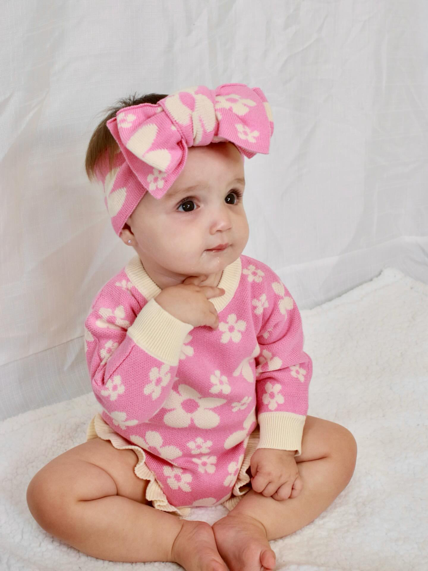 Baby wearing our Violet Knit Headband and matching pink floral knit romper sitting on a soft white backdrop
