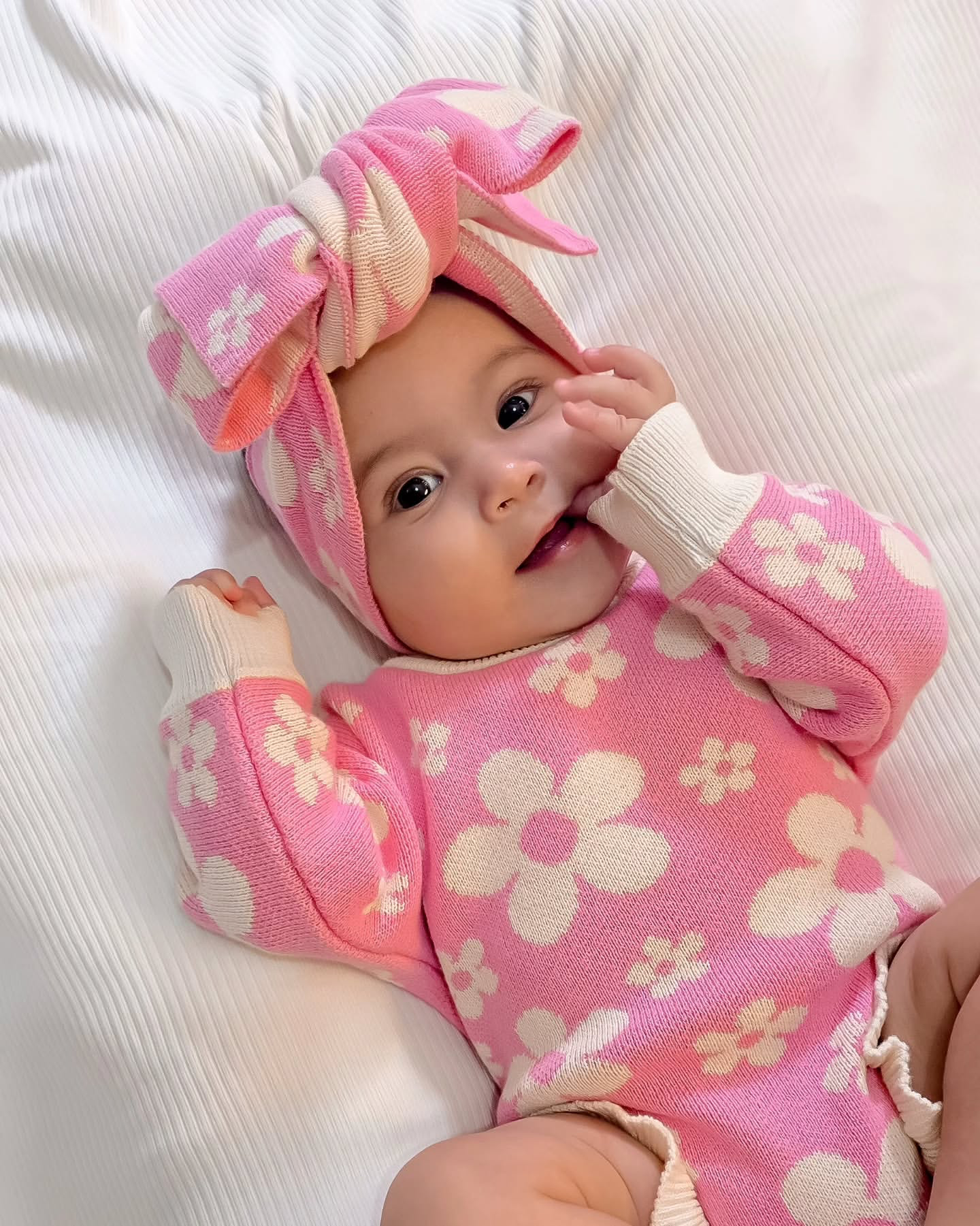Baby wearing our Violet Knit Headband and matching pink floral knit outfit lying on soft white bedding