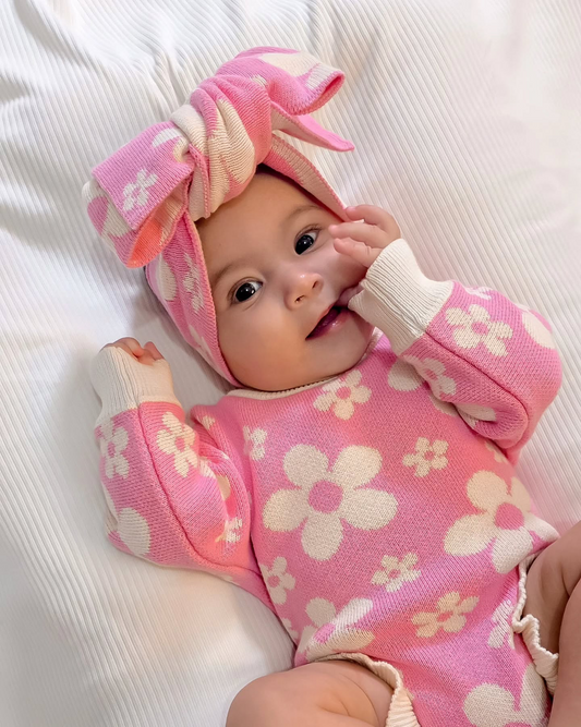 Baby wearing our Violet Knit Headband and matching pink floral knit outfit lying on soft white bedding