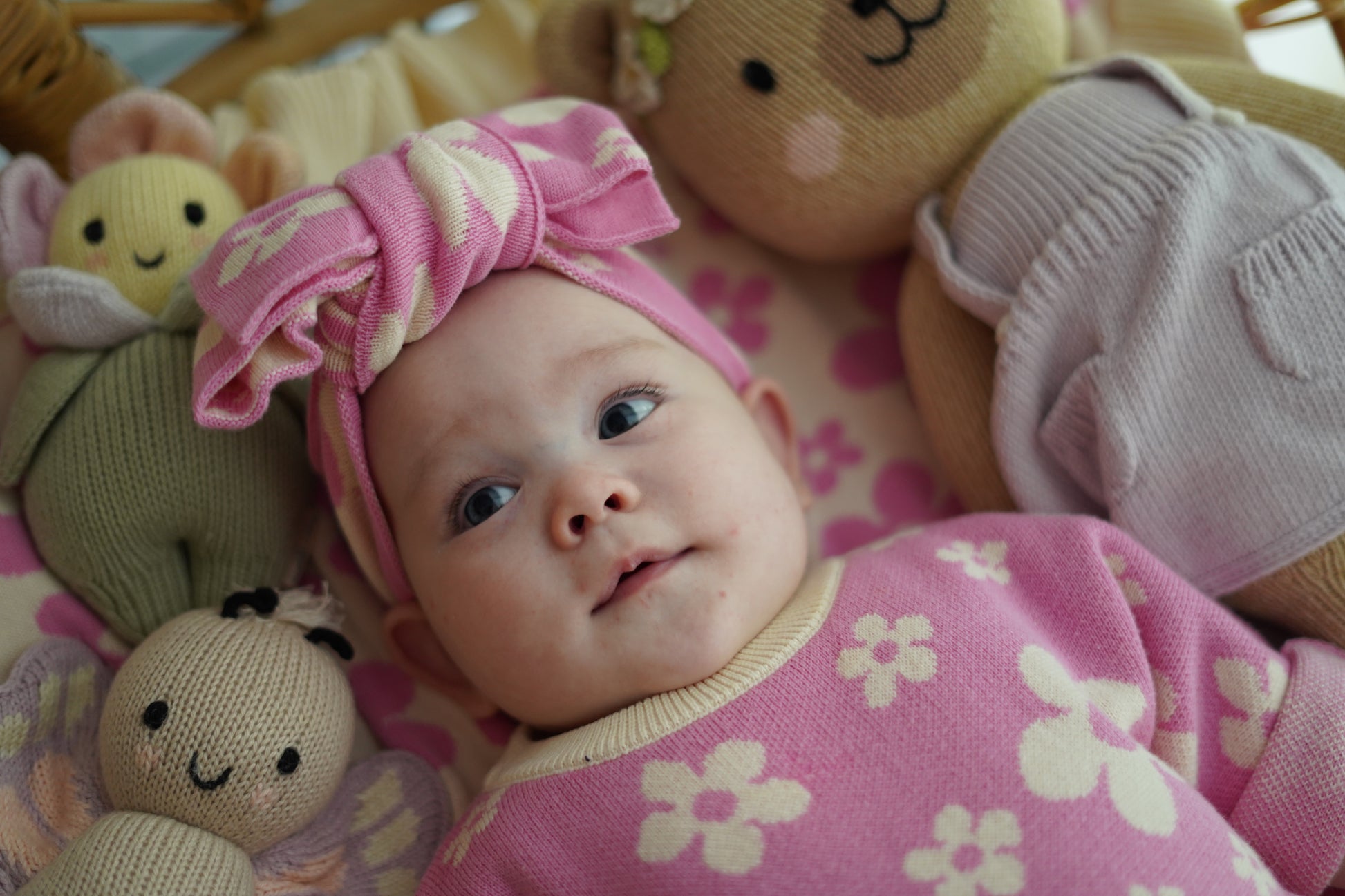 Baby wearing our Violet Knit Headband and matching pink floral knit outfit lying with soft plush toys in a cosy styled setting