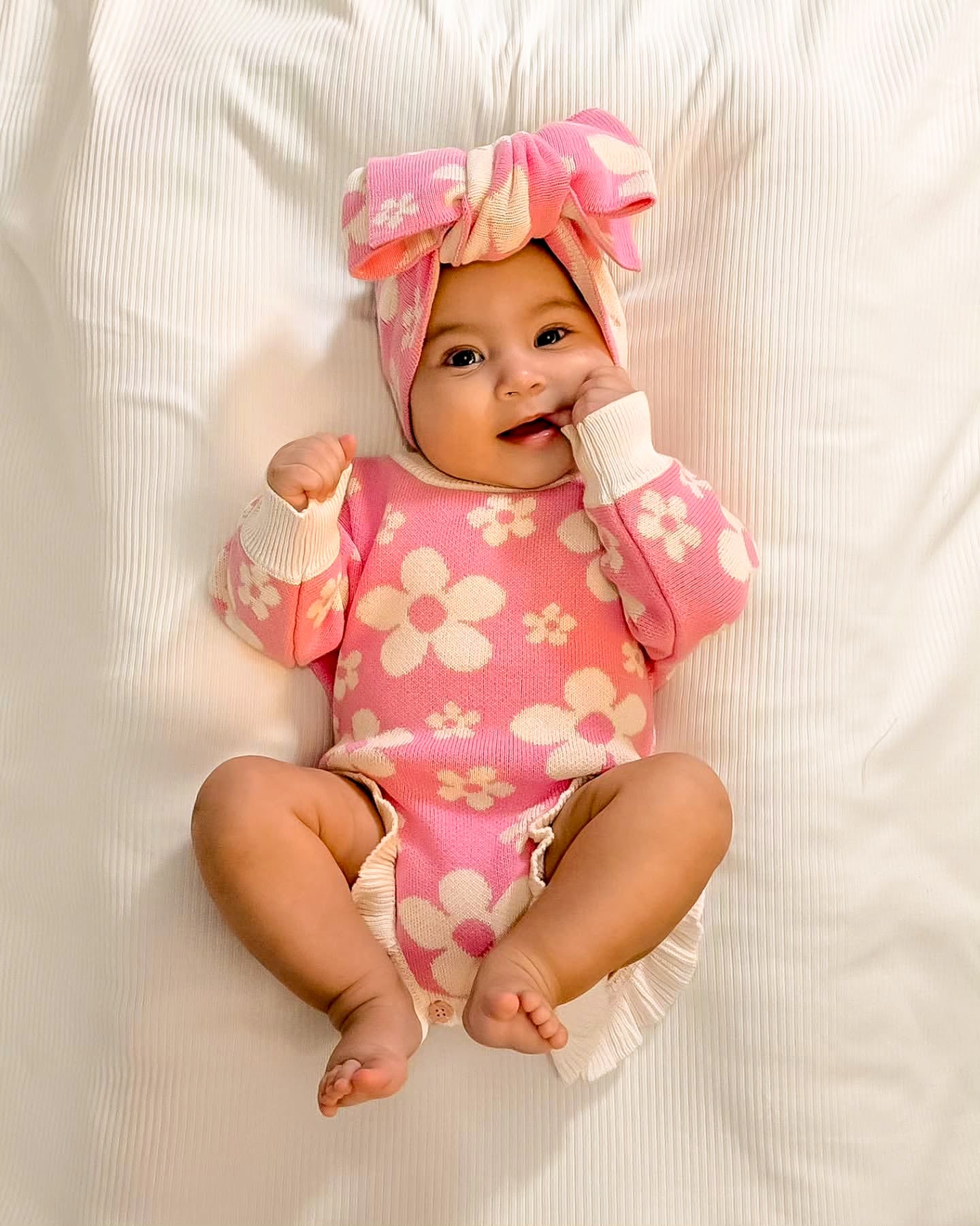 Baby wearing the Little Violet Threads Violet Ruffle Romper in pink floral knit with matching oversized bow headband, lying on white bedding
