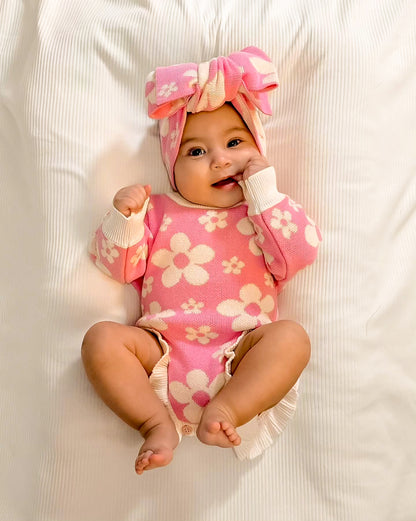 Baby wearing the Little Violet Threads Violet Ruffle Romper in pink floral knit with matching oversized bow headband, lying on white bedding