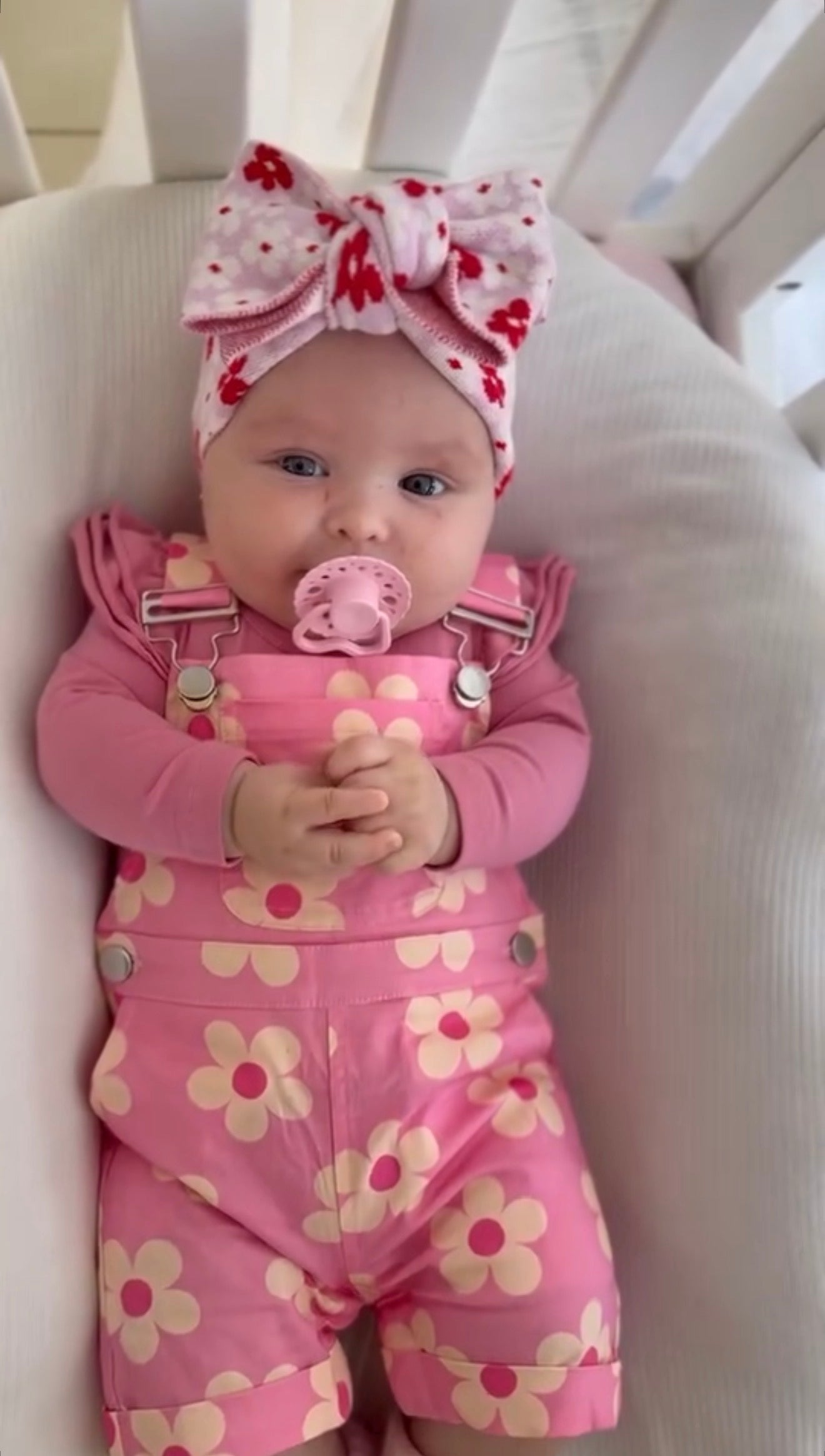 Baby in pink floral outfit with a pacifier and headband, sitting in a crib.