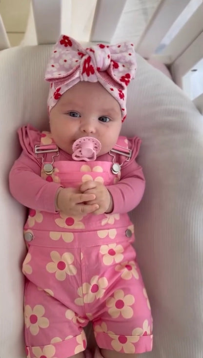 Baby in pink floral outfit with a pacifier and headband, sitting in a crib.